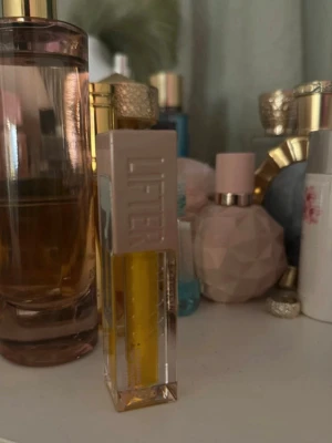 Maybelline Lifter Gloss i gult - Säljer en Maybelline Lifter Gloss med transparent gul färg. Kommer i en stilren fyrkantig plastförpackning med beige lock och tydlig logga på sidan. Perfekt för att ge läpparna en glansig finish och extra volym.