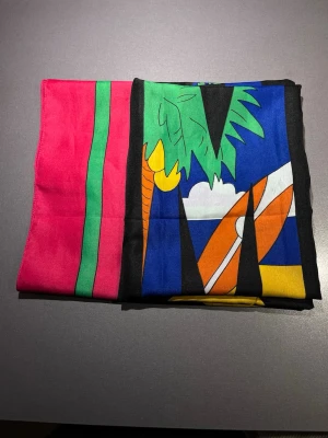 Färgglad strandduk/sjal  - Färgglad strandduk/sjal med ett stort tryck av palmer, surfbrädor och stadssilhuett. Färgerna är starka med blått, grönt, rosa, orange, svart och gult. Ett somrigt motiv som sticker ut på stranden 😎🏝️ Supermätt och bra att ha på picknicken eller stranden. Mått: 160x120cm. Mycket bra skick! Säljs på flera sidor, först till kvarn. 