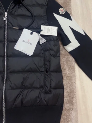 Svart cardigan från Moncler med huva - Säljer en svart dunväst från Moncler med quiltad design och huva med dragsko. Västen har en silverfärgad dragkedja och ett stort vitt mönster på ena axeln. Materialet är polyester och dun, perfekt för kyliga dagar. Moncler-logga på axeln för extra stil.