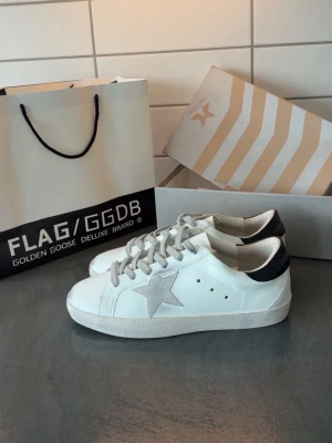 Golden Goose vita sneakers med stjärna - Säljer ett par klassiska vita Golden Goose sneakers med grå mockastjärna på sidan, grå snörning och svart hälparti i skinn. Sidan har GGDB/SSTAR-tryck och sulan är vit med struktur. Skorna är i skinn och mocka, perfekta för dig som gillar stilrena detaljer.