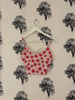 Rosa croppad linnetopp med jordgubbar - Säljer en söt rosa croppad linnetopp från SHEIN i storlek XS. Toppen har tunna axelband och är täckt med ett lekfullt jordgubbsmönster i rött. Materialet är mjukt och lätt polyester, perfekt för varma dagar. Den har vågig kant nedtill för extra detalj.