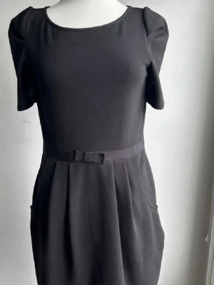 Svart kortklänning Juicy Couture - Säljer en svart kortklänning från Juicy Couture med rund halsringning, korta ärmar och en söt liten rosett i midjan. Klänningen har fickor och lätt puffade axlar för en cool siluett. Tillverkad i mjukt material som känns bekvämt hela dagen.