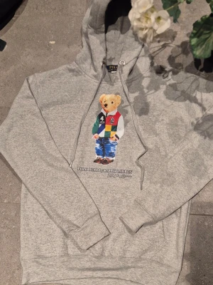 Grå polo bear ralph lauren - * 🧥 Typ: Pullover-hoodie (utan dragkedja) * ✨ Skick: Nyskick – mycket fint skick utan synligt slitage * 🎨 Färg: Mörk svart/mörkgrå, jämn och fräsch ton * 🏷️ Märke: Nike – stilren vit logotyp på vänster bröst * 🧵 Material: Mjuk och följsam bomullsblandning av hög kvalitet * 👕 Passform: Bekväm och klassisk, lätt oversized känsla * 🧢 Huva: Huva med justerbara dragsnören * 🧤 Detaljer: Praktisk magficka framtill * 👌 Stil: Tidlös och lättmatchad, perfekt till vardag och tillställningar