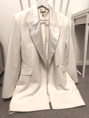 Vit kavaj från H&M med breda slag - Säljer en stilren vit kavaj från H&M med breda slag och en knapp framtill. Kavajen har två fickor med lock och är helfodrad. Perfekt för dig som vill ha en clean och klassisk look med en modern twist.