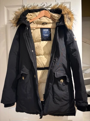 Svart parkas från Svea med fuskpäls - Säljer en svart parkas från Svea med beige foder och stor huva med fuskpäls. Jackan har dragkedja, knappar och två stora fickor framtill. På ena ärmen sitter en patch med amerikansk flagga. Perfekt för kalla dagar och riktigt snygg streetstil.