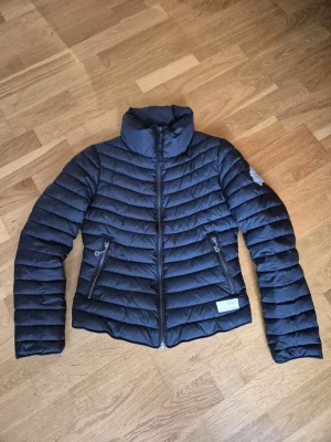 Svart pufferjacka från Odd Molly - Snygg mörkblå pufferjacka från Odd Molly med coolt emblem på ärmen och två dragkedjeförsedda fickor. Jackan har horisontella quiltade sömmar och hög krage som ger extra stilpoäng. Perfekt för dig som vill ha en trendig look när det är kallt ute.