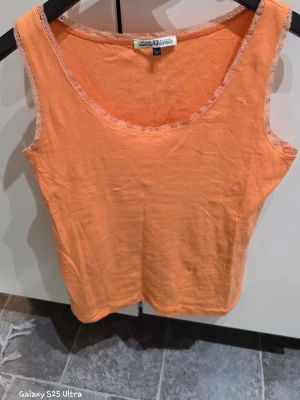 Orange linnetopp från Hampton Republic - Snygg orange linnetopp från Hampton Republic i storlek L. Toppen är ärmlös och har spetsdetaljer längs halsringningen och axelbanden. Tillverkad i mjuk bomull, perfekt för varma dagar och enkel att matcha med jeans eller shorts.