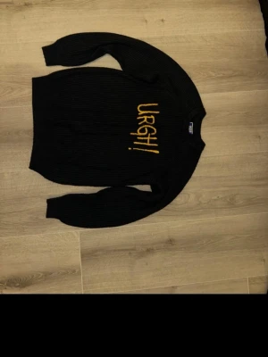 Svart stickad tröja Jack & Jones - Svart stickad tröja från Jack & Jones med ribbad struktur och trycket 'URGH!' i gul färg på framsidan. Rund hals och långa ärmar, perfekt för dig som gillar streetstyle och vill ha något som sticker ut lite extra.
