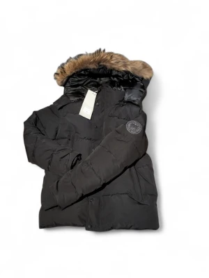 Canada Goose - Svart pufferjacka från Canada Goose med stor huva och snygg pälsdetalj. Jackan har ett broderat märke på ärmen, praktiska fickor och knappar framtill. Perfekt för kalla dagar och har en riktigt schysst look som sticker ut. Är i storleken M 175-185