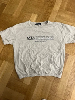 Svea sport T-shirt  - Vit t-shirt från Svea Sport Club i storlek XS med tryck på bröstet. Modellen är kort ärmad och har rund hals, samt en elastisk kant nedtill som ger en cool och avslappnad vibe. Materialet är mjuk bomull, perfekt för chill idrotten eller gymmet.