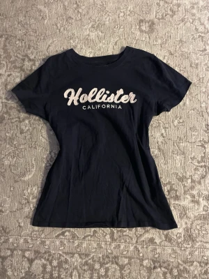 Marinblå Hollister t-shirt med tryck - Snygg marinblå t-shirt från Hollister med vit broderad logga och texten 'California' på bröstet. Modellen är klassisk med rund hals och korta ärmar, perfekt för en chill och avslappnad stil. Tillverkad i mjuk och skön bomull. Dm för mått och frågor 💘