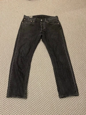 Levi's 501 grå jeans  - Svarta Levi's 501 jeans med klassisk rak passform och fem fickor. Jeansen har knappgylf och detaljer som synliga nitar och Levi's patch bak i midjan. Materialet är slitstark denim i bomull. Perfekt för dig som gillar tidlös stil och vill ha ett par jeans som funkar till allt.