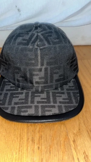 Svart mönstrad Fendi keps - Säljer en svart keps från Fendi med deras ikoniska FF-mönster över hela kepsen. Kepsen har justerbar rem baktill med Fendi-logga i guld. Materialet är polyester och bomull, vilket gör den både snygg och bekväm. Perfekt accessoar för att lyfta din streetwear-stil.