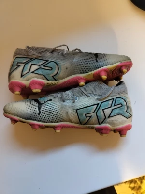 Grå/blå Puma fotbollsskor med dobbar - Säljer ett par grå fotbollsskor från Puma med snörning och svart Puma-logga på ovansidan. Skorna har blå detaljer och stora FTR-tryck på sidan. Sulan är rosa med gula dobbar, perfekt för gräsplan och konstgräs. Ovandelen är i syntetmaterial och har en rund tå.