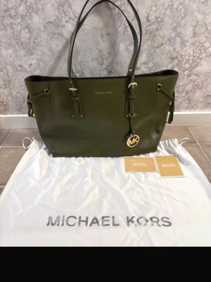 Michael Kors handväska khaki - Snygg olivgrön handväska från Michael Kors i skinn med guldfärgade detaljer och logotyp. Väskan har två långa axelremmar, flera innerfack och dragkedja. Perfekt storlek för att få plats med allt du behöver och stilren design som lyfter din look.