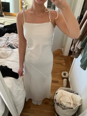 balklänning  - Säljer en stilren vit långklänning med smala axelband som knyts på axlarna. Klänningen har rak passform och ärmlös design med fyrkantig urringning. Materialet känns lätt och svalt, perfekt för varmare dagar. Enkel och elegant look som är lätt att matcha med accessoarer.