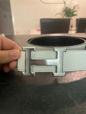 Hermmes  - Säljer en stilren väst i vitt och svart skinn med ett stort H-format metallspänne framtill. Västen har en klassisk, rak form och är tillverkad i slätt och strukturerat skinn. Perfekt för dig som gillar minimalistisk design och vill ha en cool detalj i din outfit.