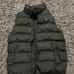 Svart dunväst från Gant - Snygg svart dunväst från Gant med dragkedja och hög krage. Perfekt för kyligare dagar och enkel att matcha med andra plagg. Västen har praktiska fickor med dragkedja för säker förvaring.