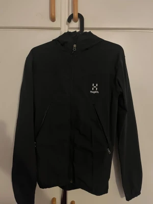 Svart vindjacka från Haglöfs - Svart vindjacka från Haglöfs med huva och hel dragkedja framtill. Jackan har två snedställda fickor med dragkedja och diskret logga på bröstet. Perfekt för utomhusaktiviteter och har en stilren, enkel design.