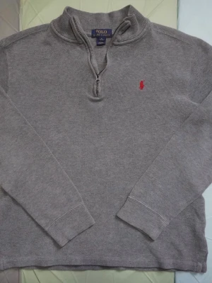 Grå stickad tröja Polo Ralph Lauren - Snygg grå stickad tröja från Polo Ralph Lauren i storlek XL men sitter som L. Tröjan har en half zip-dragkedja vid halsen och ett rött broderat Polo-logo på bröstet. Långärmad med ribbade muddar och klassisk, enkel design som funkar till många stilar.