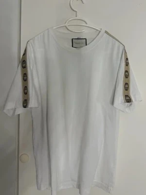 Gucci  - Säljer en clean vit t-shirt från Guccikk med korta ärmar och beige band med svarta Gucci-loggor längs axlarna. T-shirten har en klassisk rund hals och är gjord i mjuk bomull som känns skön mot huden. Perfekt för dig som gillar stilrena designer med lyxig touch.