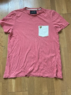 Rosa t-shirt från Lyle & Scott - Snygg rosa t-shirt från Lyle & Scott med vit bröstficka och gul broderad logga. T-shirten har rund hals och korta ärmar, perfekt för en chill stil. Materialet är mjuk bomull som känns skönt mot huden.