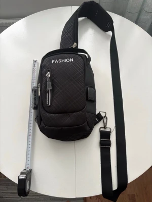 Svart axelväska med rutigt mönster - Säljer en svart axelväska från FASHION med rutigt mönster och vita detaljer. Väskan har flera dragkedjor, USB-port och hörlursuttag på sidan samt justerbar rem. Materialet är slitstark polyester och designen är modern och praktisk för vardagsbruk.