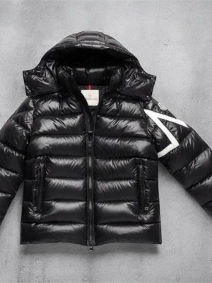 Svart pufferjacka från Moncler - Säljer en svart pufferjacka från Moncler med glansig finish och stor huva. Jackan har en vit broderad detalj på ena ärmen, två fickor med dragkedja och tydliga quiltade sektioner. Perfekt för dig som vill ha en snygg och varm jacka med streetkänsla.