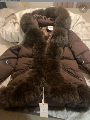 Brun pufferjacka med päls från Copperrose - Brun pufferjacka från Copperrose med fluffiga pälsdetaljer längs huva, krage och nederkant. Jackan har bälte i midjan och dragkedja framtill. Perfekt för kalla dagar när du vill vara både varm och snygg. Pälsen är mörkbrun och jackan har en oversized känsla.