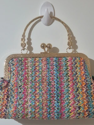 Färgglad handväska med gulddetaljer - Unik handväska med flätat, flerfärgat syntetmaterial och snygga guldfärgade metalldetaljer. Väskan har ett klassiskt handtag och ram med dekorativa mönster, samt ett svart innerfoder. Perfekt för dig som gillar färg och vill sticka ut.🌈🛍😍