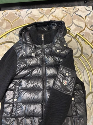 Svart dunväst från Moncler med huva - Säljer en svart dunväst från Moncler med stickade sidor och rygg, quiltad framsida och avtagbar huva. Västen har dragkedja framtill, två fickor med tryckknappar och en snygg Moncler-logga på ärmen. Perfekt för dig som gillar exklusiva limited cardigans. Funkar för alla års tider, verkligen en sjukt snygg cardigan.