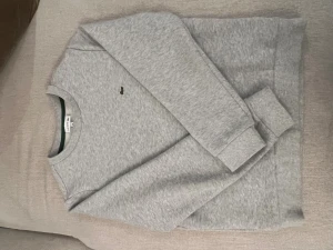 Grå sweatshirt från Lacoste i barn storlek  - Säljer en stilren grå sweatshirt från Lacoste med den klassiska gröna krokodilloggan på bröstet. Tröjan har rund halsringning, ribbade muddar och är tillverkad i mjuk bomull. Perfekt för dig som gillar enkel och clean stil.