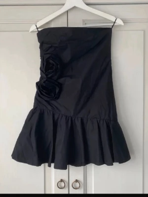 Svart axelbandslös kortklänning med rosdetaljer - Så fin axelbandslös svart kort klänning med snygga detaljer!! Endast använd någon gång. Perfekt till nyår, jag skickat snabbt! Pris kan diskuteras vid snabb affär 