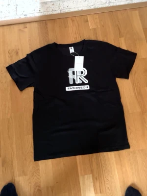 Svart t-shirt med Fashweon-tryck - Svart t-shirt från GUEEN med stort grafiskt Fashweon-tryck i vitt och grått på bröstet. Rund hals och korta ärmar. Materialet är mjuk bomull som känns skönt mot huden. Perfekt för dig som gillar streetwear och tydliga prints.