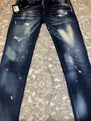Mörkblå jeans med färgstänk från Dsquared2 - Säljer ett par mörkblå jeans från Dsquared2 med coola vita färgstänk och slitningar på framsidan. Baktill finns textbroderi på ena fickan och tydlig Dsquared2-tag. Jeansen har klassisk femficksdesign och är tillverkade i jeansmaterial.