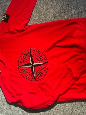 Röd sweatshirt från Stone Island - Säljer en röd långärmad sweatshirt från Stone Island i bomull. Tröjan har ett stort broderat Stone Island-emblem på bröstet och den klassiska patchen på ärmen. Rund hals och normal passform, perfekt för dig som gillar streetwear.