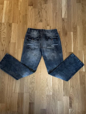 Tom Tompson Bootcut jeans - Svarta jeans från Tom Tompson med coolt tvättad look och kontrastsömmar. Modellen är Bootcut och har de klassiska fickor med knappstängning bak. Materialet är slitstarkt jeans och midjan är normal. Perfekt för dig som gillar en unik och stilig look. 