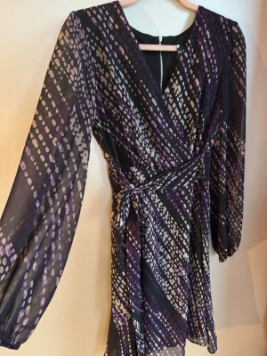 Svart och lila chiffongklänning - Snygg midiklänning från Maison Tara med svart bas och lila samt beige prickigt mönster. Klänningen har långärmade, transparenta ärmar och knyt i midjan. V-ringad framtill och dragkedja i ryggen. Fint chiffongmaterial som ger en elegant look.