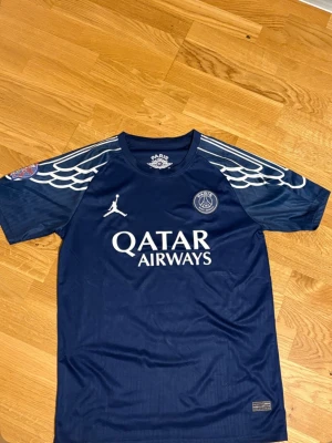 PSG Jordan special edition tröja – Navy – Storlek M (passar S) - Exklusiv PSG x Jordan special edition-tröja i marinblått. Ikoniska vita vingdetaljer på axlarna, Jordan-logga, PSG-klubbmärke och Qatar Airways-tryck på framsidan. Tillverkad i lätt och andningsbart sportmaterial som sitter bekvämt vid träning och vardag.  Storlek står som L, men känns mer som M/S i passform. Mycket fint skick utan skador eller fläckar. Perfekt för dig som älskar PSG eller Jordan-kollektionerna