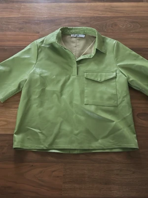 Grön skinnskjorta från NA-KD - Cool grön skjorta i skinnimitation från NA-KD, med korta ärmar och klassisk krage. Framsidan har en stor ficka med lock och baksidan har knappar hela vägen. Perfekt för dig som gillar statement-plagg och vill sticka ut med en unik färg. Köptes second hand.