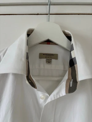Vit Burberry skjorta med rutiga detaljer - Säljer en vit skjorta från Burberry i slim fit med klassiska rutiga detaljer på krage och manschetter. Skjortan har långa ärmar, knappar framtill och ett litet Burberry-emblem broderat på bröstet. Materialet är mjuk bomull och passformen är normal.