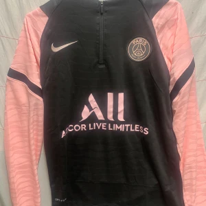 PSG långärmad träningströja Nike M - Paris Saint-Germain långärmad träningströja från Nike i svart med ljusrosa detaljer och klubbmärke på bröstet. Tröjan har halv dragkedja, Dri-FIT-material och snygga mönstrade ärmar. Perfekt för dig som älskar fotboll och vill sticka ut på planen.