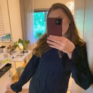 Mörkblå blus med volangdetaljer - Säljer en mörkblå blus med långärmade ärmar och volangdetaljer vid halsringning och ärmslut. Blusen har en lätt strukturerad yta och en avslappnad passform, perfekt för dig som gillar stilrena men ändå unika plagg. Aldrig använd, har bara legat i garderoben, är villig att stryka den innan postningen🤎🤎