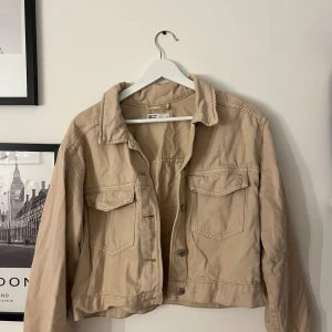 Beige jeansjacka från Gina Tricot - Snygg beige jeansjacka från Gina Tricot med klassisk krage, två bröstfickor med lock och knappar framtill. Jackan är croppad och har långa ärmar, perfekt för en trendig och avslappnad look.