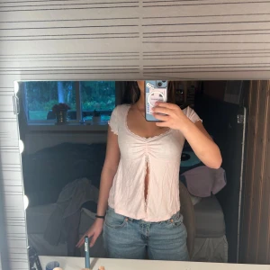 Ljusrosa blus från Gina Tricot - Supersöt ljusrosa blus från Gina Tricot med korta ärmar och spetsdetaljer vid halsringningen. Toppen har en snygg v-ringad front med snörning och en liten slits framtill. Perfekt för dig som gillar romantisk och feminin stil.