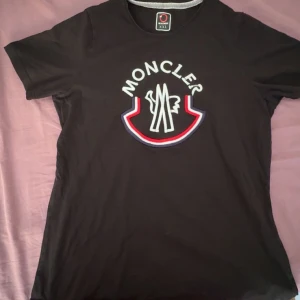 Svart Moncler t-shirt med broderad logo - Svart t-shirt från Moncler i bomull med stor broderad logotyp framtill i vitt, rött och blått. Klassisk rund hals och korta ärmar. Perfekt för dig som gillar streetwear och vill sticka ut med en fet logga.