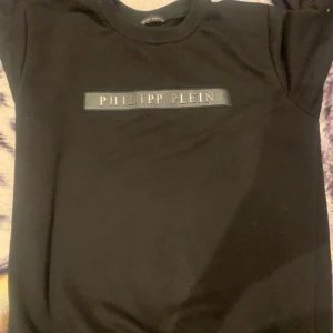 Svart Philipp Plein sweatshirt - Svart sweatshirt från Philipp Plein med klassisk rund hals och silverfärgat logotyptryck framtill. Tröjan har ribbade muddar och en broderad PP-logga på ärmen. Tillverkad i mjuk bomull för en skön och stilren look.