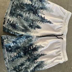 Vita meshshorts med blått skogsmotiv - Säljer ett par vita meshshorts i storlek M med blått och mörkgrönt skogsmotiv. Shortsen har elastisk midja och svart snörning framtill. Tillverkade i 100% polyester, perfekta för sommardagar eller träning.