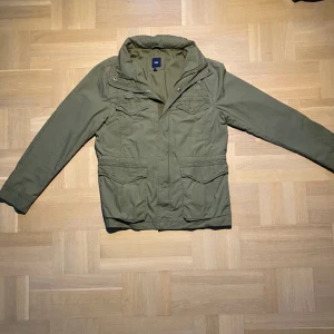 Grön Field jacket - Jag säljer en riktigt snygg field jacka som är perfekt nu inför hösten! Jackan har en luva som man kan öppna med hjälp av dragkedja🙌 Jackan passar dunder med jeansen som du hittar i min profil!! Tveka inte på att ställa frågor😁
