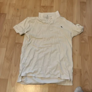 Vit pikétröja från H&M - Klassisk vit pikétröja från H&M med korta ärmar och krage. Tröjan har knäppning framtill och en liten broderad logga på bröstet. Tillverkad i mjuk bomull, perfekt för en clean och enkel stil.